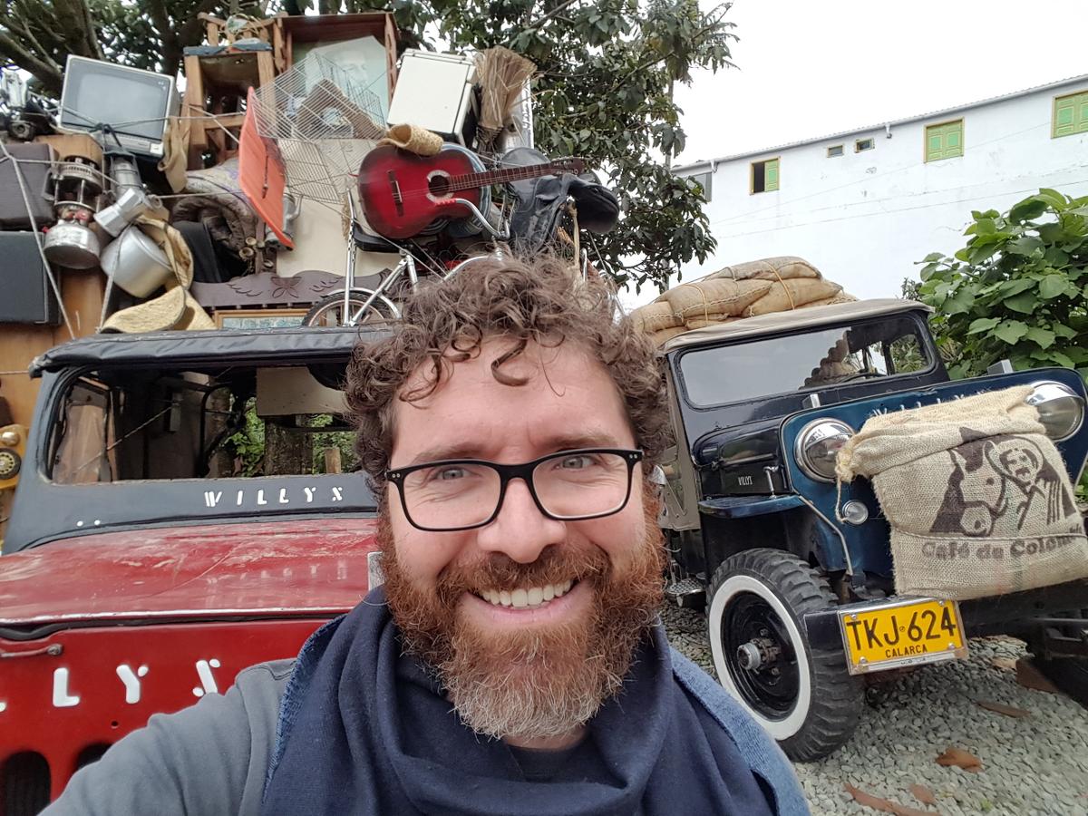 Photo of Francisco Posada standing in front of vehicles, stacked high with items, with Colombian license plates.